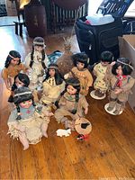 Photo of seven Native American style dolls arranged around a small teepee on wooden floor, various sitting and standing poses with traditional clothing.