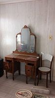 Full view of wood vanity with three-panel mirror and wood chair with floral upholstery next to it