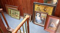 Group of framed wall art pieces leaning against a wall along a stair landing featuring portraits and landscapes.
