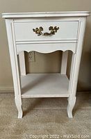 Front angle view of white accent table showing drawer with ornate handle and lower shelf.