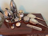 Wide view of all lot items on dark wood surface including silver mirror, brush, comb, two vanity jars, two reflective vases, and tray