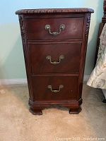 Front view of the small wooden cabinet showing three drawers with ornate metal handles and carved decorative edges.