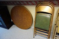 One photo showing a round card table leaned against a wall and six folded chairs propped next to it, displaying the brown table surface and the green vinyl on chair seats and backs.
