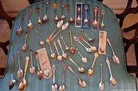 Full collection of 31 assorted souvenir spoons laid out on striped fabric surface, showing variety of styles and designs.