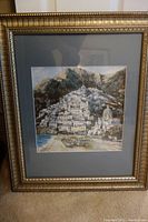 Full view of framed art depicting a coastal village landscape with boats and white buildings on a hillside framed in a gold-colored decorative frame with minor scratches.