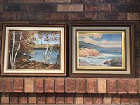 Two framed paintings side by side showing landscapes with water, trees, and rocky coast. Frames have bronze and gold accents.