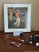 Framed golf-themed art above fishing rods, reel, fishing line box, and lures on floor