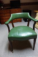 Front view of a green leather armchair with wooden frame and casters on legs, showing curved back and armrests with nailhead trim.