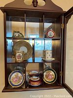 Close-up of the wooden wall mount display cabinet showing porcelain items inside, including floral and romantic scene decorated plates and cups