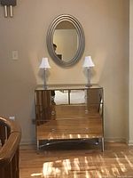 Mirrored chest cabinet with two silver lamps and oval mirror mounted on wall above.