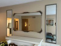 Three wall mirrors displayed above a white mantel. Largest mirror has ornate corner decorations. Two narrow mirrors flank the large mirror.