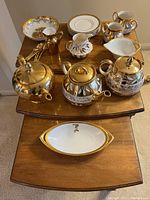 Overview of table with three ornate tea pots, gold-toned pitcher, floral dish, multiple small cups and plates, and the large white and gold monogrammed serving dish on pull-out table.