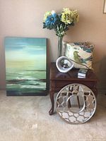 View of abstract painting, round silver decorative mirror, floral vase, bird throw pillow, and small decorative boxes placed on wooden table next to painting leaning against wall