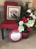 Red chair with framed floral artwork resting on seat, round red leather-framed mirror on floor, large ceramic vase with artificial flowers nearby.
