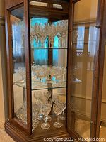 Three shelves of crystal and stemware glasses stored in a curio cabinet, displaying varied wine glasses and goblets on glass shelves.