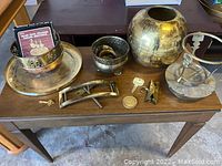Photo displaying the full lot of miscellaneous yellow brass items arranged on a wooden table including vase, candle holder, keys, whistle, buckle and a booklet.