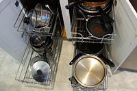 View of various used stainless steel pots, non-stick pans including a Tefal pan, and lids arranged on kitchen wire shelving.