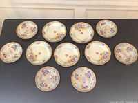 13 hand-painted Royal Vienna porcelain plates with purple and white floral designs and gold scalloped edges laid out on a black surface.