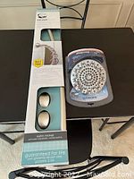 Photo showing the satin nickel curtain rod in retail packaging with two brackets and Waterpik Rainfall Optiflow shower head in packaging on a table.