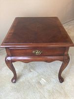 Front angle view of wood side table showing drawer with brass handle and curved cabriole legs on carpeted floor.