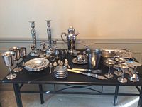 Full view of mixed silver plated set on black table, including candlesticks, goblets, serving utensils, cocktail shaker, small bowls, trays, and ice bucket.