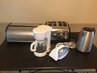 Photo showing metal breadbox, white drip coffee maker, four-slice stainless steel toaster, white clothes iron, and stainless steel electric kettle on a table.