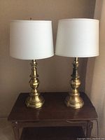 Pair of two metal table lamps with white drum shades on a wooden surface against a neutral wall. Lamps have a brass-like finish with some tarnish and show signs of wear.