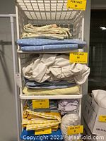 Stacked folded bed linens including sheets, mattress covers, and pillow on three top shelves of a white plastic shelf.