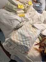 Pile of assorted bedding items including blankets and sheets with yellow 'For Sale' labels, featuring a queen size duvet cover with striped pattern