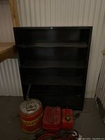 Front view of the black metal shelf showing three shelves and its back panel inside a storage or garage space. Red and silver gasoline containers are placed in front of it.