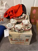 Four-piece Christmas Village ironstone place setting including teacups and plates with Christmas-themed designs placed on a box labeled for oranges.