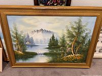Full front view of one framed painting illustrating the landscape scene with mountains, lake, waterfall, trees, and a small building in a gold-toned frame.