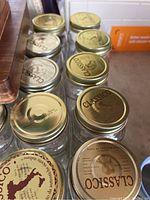 Multiple clear glass Mason jars with gold-colored metal lids printed with 'Classico' brand name, arranged on kitchen counter.