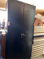 Front view of black metal storage cabinet with two doors and locking handle. Cabinet is tall with the lock centered between the doors.