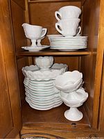 Milk glass cups stacked on saucers and plates. Several dessert bowls with grape pattern neatly stacked. Two milk glass pedestal candleholders at front.