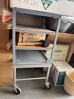 Full view of rolling cart with three particle board shelves and metal frame. Cart is filled with boxes and a mallet, which are not included.