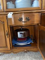 Wooden hutch lower cabinet showing stainless steel cake cover with glass plate, blue cloth underneath, and pewter candlestick partially visible next to serving pieces on bottom shelf.