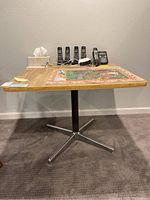 Alternate angle of square laminate table with metal base on carpeted floor, showing four phones and other items on top for scale (items not included).