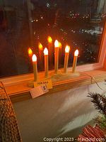 Seven white vintage electric candle lights with orange flame bulbs emitting a warm glow on a white base by a window.