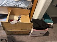 Two cardboard boxes containing folded women's clothing items, various colors and fabrics visible including black pants and light fabrics.