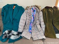 Three women's coats on sofa with scarves: teal Ashley Scott wool coat with fuzzy scarf, beige puffer coat with fur-trimmed hood and multicolor scarf, green suede coat with fur collar.