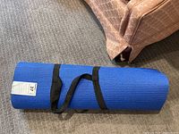 Rolled blue yoga mat secured with black carrying strap, placed on carpet.