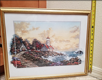 Photo of a coastal lighthouse art print in a gold frame showing detailed scene including buildings, rocky shoreline, sea and sky, with a measuring tape indicating about 20 inches width.