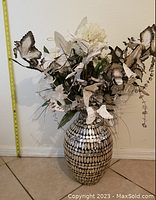 Full view of tall vase with intricate black and white shell mosaic pattern holding artificial floral arrangement with butterflies and greenery.
