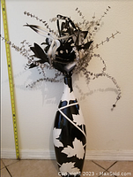 Photo of the tall black and white vase with a leaf pattern and artificial flower arrangement against a plain wall with a measuring tape beside it.