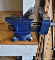 Side angle photo of blue metal workshop vice mounted on wooden surface showing serrated jaws and handle.