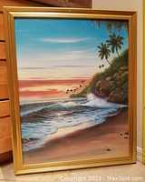 Photo of framed beach painting showing ocean waves, palm trees, and sunset in golden colored frame.