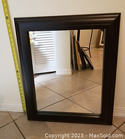Rectangular mirror with black-brown wooden frame leaning against wall, with measuring tape for scale.