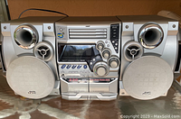 Front view of silver JVC HiFi stereo system with dual speakers, showing CD player, cassette deck, control knobs, and speakers with fabric covers.