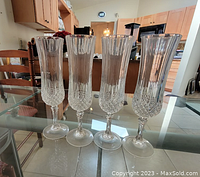 Four clear crystal champagne flutes with detailed lower half etched designs on a glass table indoors near kitchen furniture.